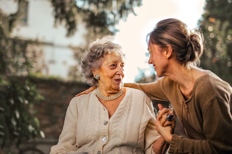 aging parents and caregiver burnout. younger woman speaking to an older woman outside surrounded by foliage.